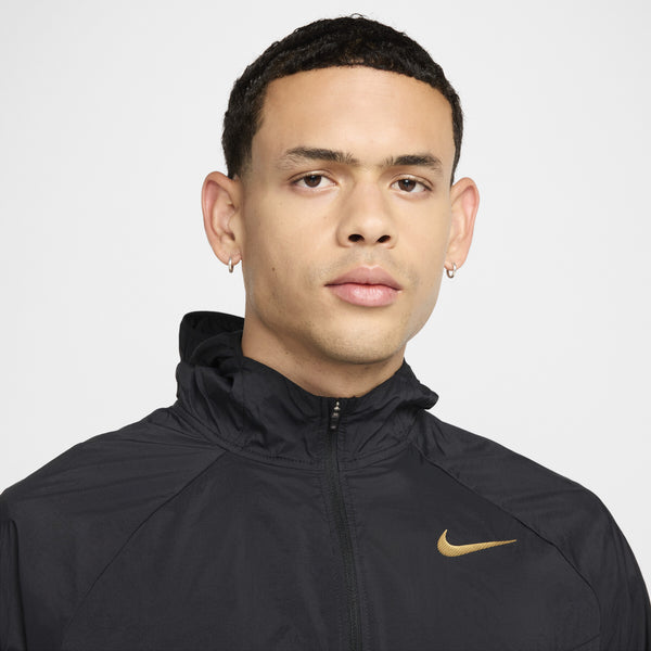 En person med kort mørkt hår, iført Nike's Nk Rpl Jakob Jkt herre i Black/Black/Metallic Gold med guld swoosh og små hoop øreringe, poserer foran en almindelig lys baggrund. Vandafvisende Dri-FIT-teknologi sikrer komfort og stil.