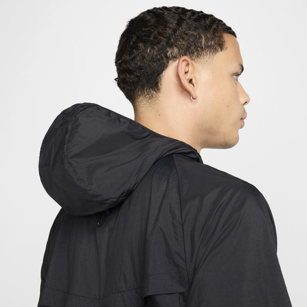 En person med kort, krøllet hår og en lille ørering ses bagfra og fra siden iført en Nike Nk Rpl Jakob Jkt, Herre, Black/Black/Metallic Gold. Hætten er oppe, men ikke over hovedet. Baggrunden er ensfarvet og lys.