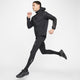 En person bærer en Nike Nk Rpl Jakob Jkt, Herre, Black/Black/Metallic Gold, sammen med sorte leggings og sneakers, fanget midt i gangen mod en hvid baggrund, hvor det ser ud til at løbe eller hoppe.