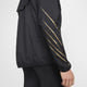 En person set fra siden er iført Nike Nk Rpl Jakob Jkt, Herre, Black/Black/Metallic Gold med fire tynde diagonale guldstriber på ærmet og sorte bukser på en ensfarvet baggrund. Kun armen og en del af overkroppen er synlig.