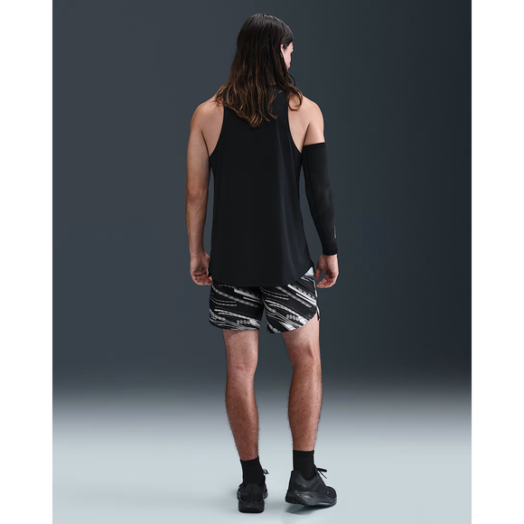 En person med langt hår står med ryggen til og er iført en sort ærmeløs top, Nike Stride 7" Brief-Lined Running Shorts i Photon Dust/Reflective Silver, sorte sokker, sorte sportssko og et sort ærme på venstre arm. Baggrunden er hvid.