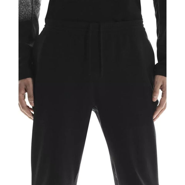 En person vises fra taljen til midt på låret iført On Weather Winter Pants, Herre, Black, og en sort langærmet skjorte med hænderne afslappet i siden. Bukserne har en elastisk linning og en løbegang.