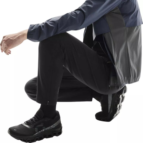 En person iført en marineblå og grå jakke, On Weather Winter Pants Herre i sort og sorte sportssko sidder på hug med venstre arm på knæet mod en almindelig hvid baggrund.