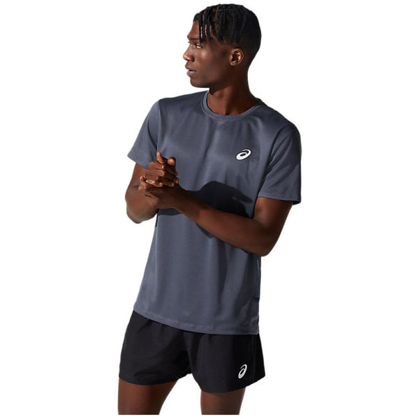 En person står foran en hvid baggrund iført en grå Asics Core SS Top, Herre, Carrier Grey løbetrøje og sorte shorts, hvor begge dele har et lille hvidt Asics-logo. Personen kigger til siden med hænderne foldet sammen.