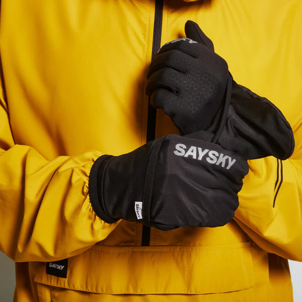 En person i gul jakke justerer sorte Saysky Pace Gloves, der har et konvertibelt luffedesign og er kompatible med touchskærme, med det hvide SAYSKY-logo synligt på bagsiden. Fokus er på handskerne og hænderne.