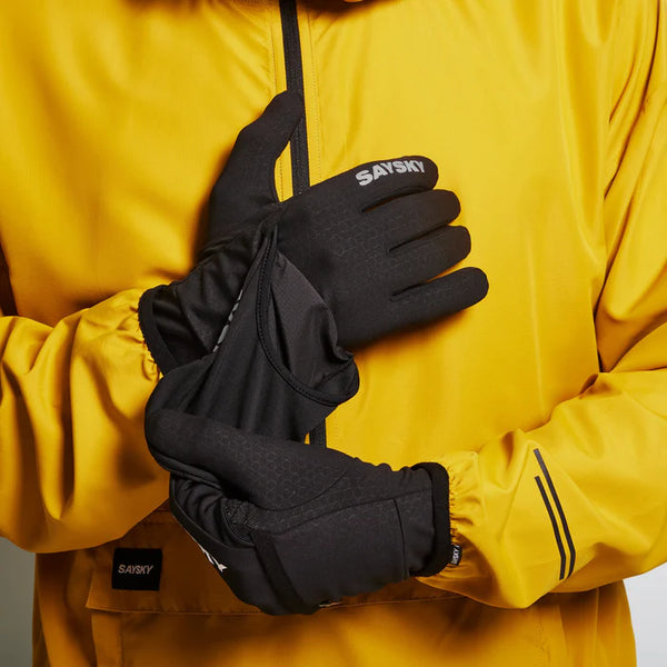 En person i en gul jakke justerer sorte Pace-handsker fra Saysky med SAYSKY-branding på ryggen og touchscreen-kompatibilitet. Den neutrale baggrund fremhæver unisex-handskerne.