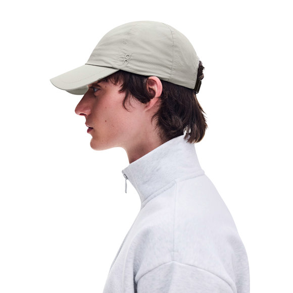 En person set i profil bærer en ON Performance Cap, Unisex, Brume, samt en lysegrå jakke med høj krave foran en ensfarvet hvid baggrund.