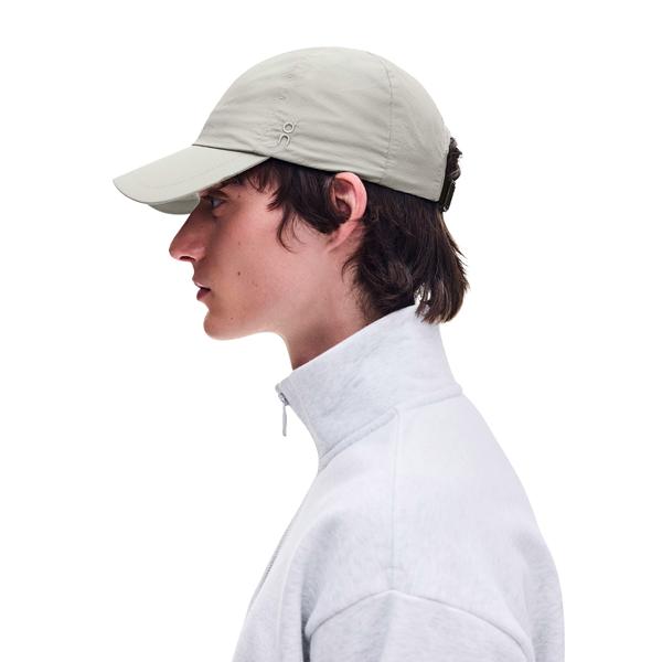 En person set i profil bærer en ON Performance Cap, Unisex, Brume, samt en lysegrå jakke med høj krave foran en ensfarvet hvid baggrund.