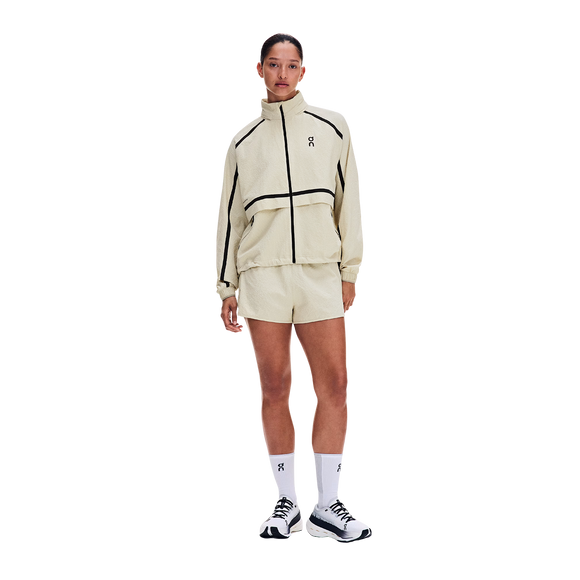 En person står foran en hvid baggrund iført ON Performance Volt-jakken, Dame, Hare, i lys beige med sorte detaljer og et lille logo, kombineret med matchende shorts og hvide sneakers. Outfittet er stilfuldt og vandafvisende.