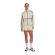 En person står foran en hvid baggrund iført ON Performance Volt-jakken, Dame, Hare, i lys beige med sorte detaljer og et lille logo, kombineret med matchende shorts og hvide sneakers. Outfittet er stilfuldt og vandafvisende.