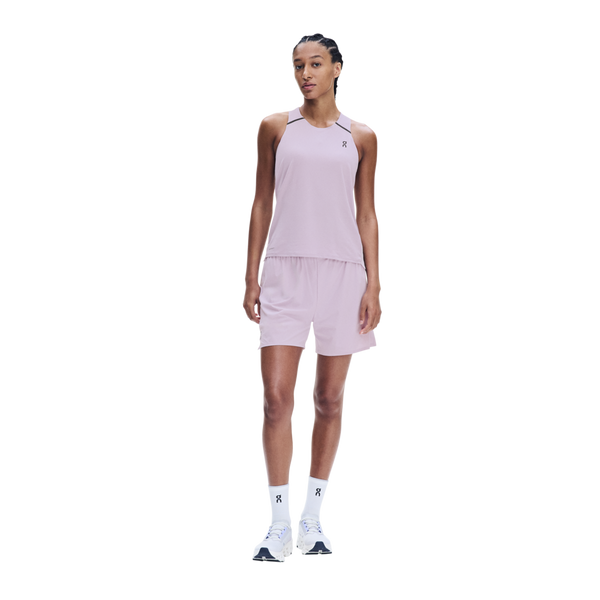 En person står med ansigtet vendt fremad iført en On Performance Tank, modellen »Dame« i farven »Foxglove/Orchid«, med matchende shorts, hvide sokker og hvide sportssko, foran en ensfarvet hvid baggrund.