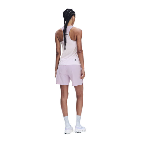 En person med flettet hår står med ryggen til kameraet og er iført en On Performance Tank, modellen »Dame« i farven »Foxglove/Orchid«, matchende shorts, hvide sokker og hvide sportssko foran en ensfarvet hvid baggrund.