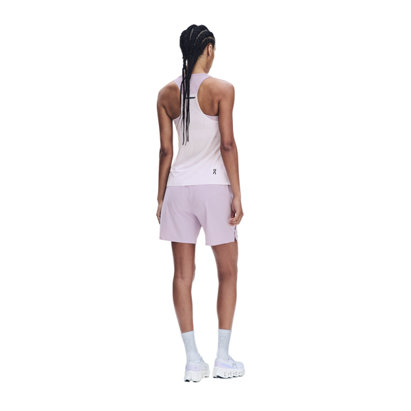 En person med flettet hår står med ryggen til kameraet og er iført en On Performance Tank, modellen »Dame« i farven »Foxglove/Orchid«, matchende shorts, hvide sokker og hvide sportssko foran en ensfarvet hvid baggrund.