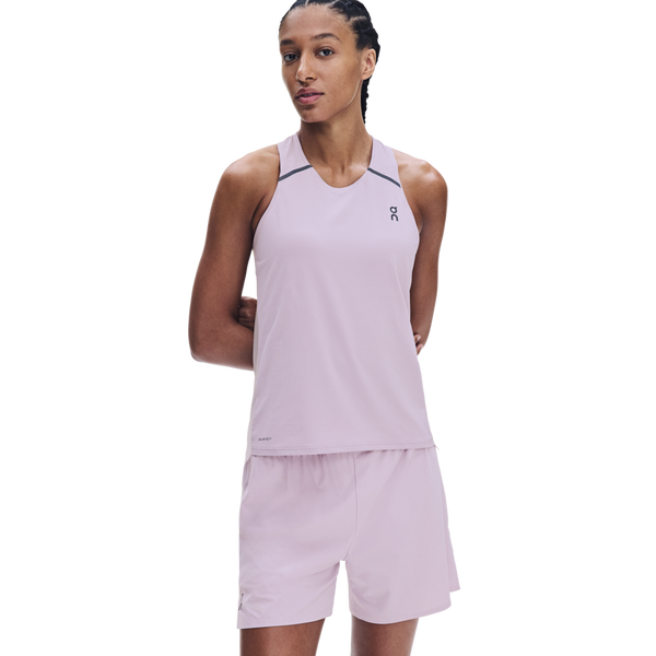 En person står med armene bag ryggen iført On Performance Tank til kvinder i farven Foxglove/Orchid, kombineret med matchende shorts designet til løb. Outfittet har diskret logo på både brystet og shortsene og er fremstillet af hurtigtørrende stof, der sikrer din komfort. Baggrunden er ensfarvet hvid.