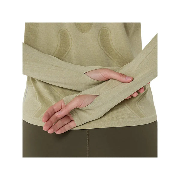 En person, der kun viser sin overkrop og sine arme, er iført Asics Road Seamless Ls Top til kvinder i Khaki/Cream med huller til tommelfingrene, parret med olivengrønne bukser og hænderne bag ryggen.