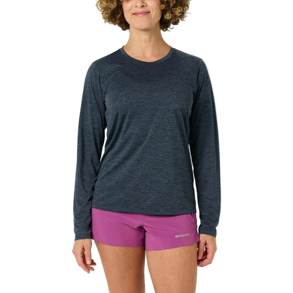 En person står med front mod en almindelig hvid baggrund iført en Patagonia Long-Sleeved Capilene Cool Daily Shirt til kvinder i Smolder Blue - Light Smolder Blue X-Dye og magentafarvede Patagonia-shorts.