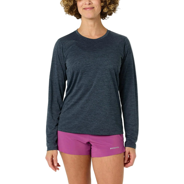 En person står med front mod en almindelig hvid baggrund iført en Patagonia Long-Sleeved Capilene Cool Daily Shirt til kvinder i Smolder Blue - Light Smolder Blue X-Dye og magentafarvede Patagonia-shorts.