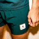 Nærbillede af en person iført Saysky Pace Shorts 5'' til mænd i grøn med et hvidt mærke med en stjerne og "SAYSKY" på venstre ben. Disse løbeshorts er lavet af åndbart materiale og er ideelle til aktive dage. Personens højre hånd er ved siden af shortsene.