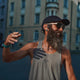 En mand med langt skæg, iført en kasket, solbriller og en tanktop, står udendørs og smiler, mens han gestikulerer med hånden. Han har Suunto Wing 2 Black Open-ear-hovedtelefoner til lyd via knogleduktion på, mens der i den slørede baggrund kan ses bygninger.