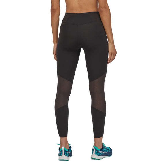 En person står med ryggen til kameraet og afslappede arme iført Patagonia Endless Run Tights til kvinder i sorte og blågrønne løbesko.