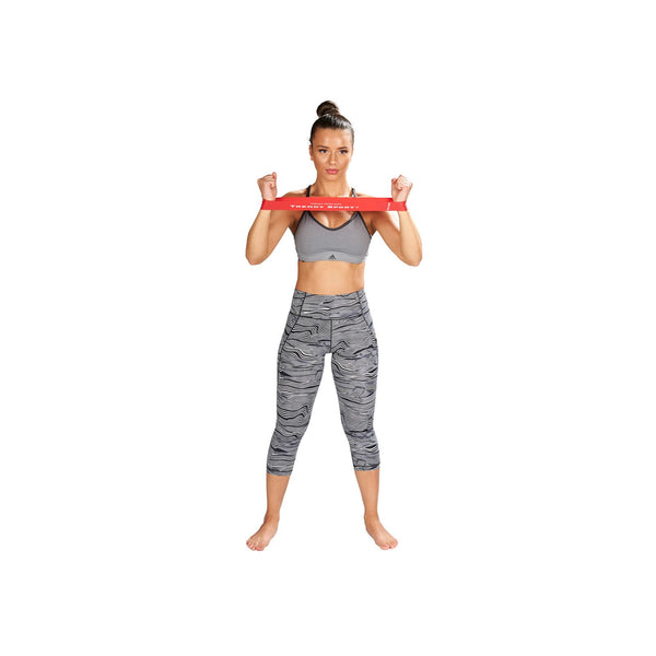 En kvinde står barfodet med spredte fødder, iført grå sports-bh og mønstrede leggings, og holder et Sports Pharma Elastikbånd, Unisex, Rød spændt i brysthøjde - ideelt til genoptræningsøvelser.
