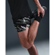 En person iført Nike Nk Df Stride 7In Bf Shrt Op2 herre løbeshorts i Photon Dust/Reflective Silver, sort skjorte og sorte sko strækker sig ved at holde den ene fod bag sig mod en almindelig, svagt oplyst baggrund.