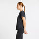 En rødhåret kvinde i profil står på en ensfarvet baggrund iført New Balance Race Day Ultra Light T-Shirt til kvinder i Grey Matter og sorte leggings med armene løst samlet bag ryggen.